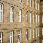 Façade massive en blocs de pierre de taille beige d'un bâtiment classique. On observe de nombreuses fenêtres blanches à petits carreaux, alignées de manière symétrique et répétitive sur plusieurs étages.