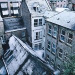 Une vue plongeante en angle élevé sur des bâtiments urbains en briques sous une fine couche de neige hivernale. On distingue plusieurs toits en pente recouverts de givre blanc, entourant une petite cour intérieure étroite. L'ambiance est calme et froide, avec des tons gris, bleus et blancs dominants.