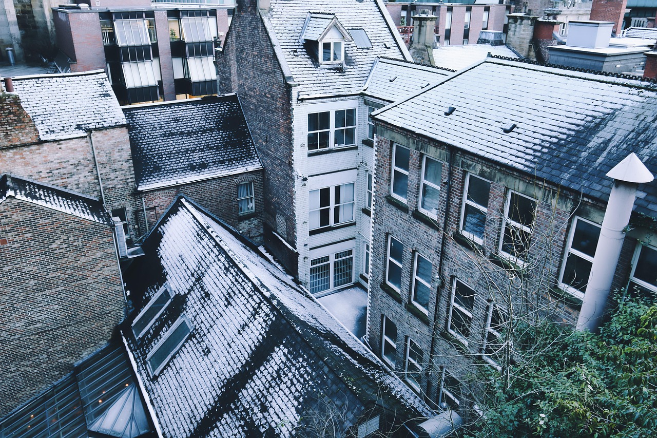 Une vue plongeante en angle élevé sur des bâtiments urbains en briques sous une fine couche de neige hivernale. On distingue plusieurs toits en pente recouverts de givre blanc, entourant une petite cour intérieure étroite. L'ambiance est calme et froide, avec des tons gris, bleus et blancs dominants.